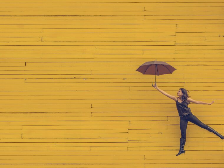 Happy woman jumping with umbrella against a sunny yellow background.